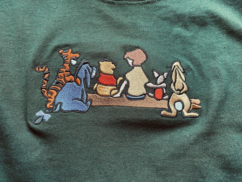 May include: Green t-shirt with an embroidered image of Winnie the Pooh and his friends, including Tigger, Piglet, Eeyore, and Rabbit. The characters are sitting on a log and smiling.