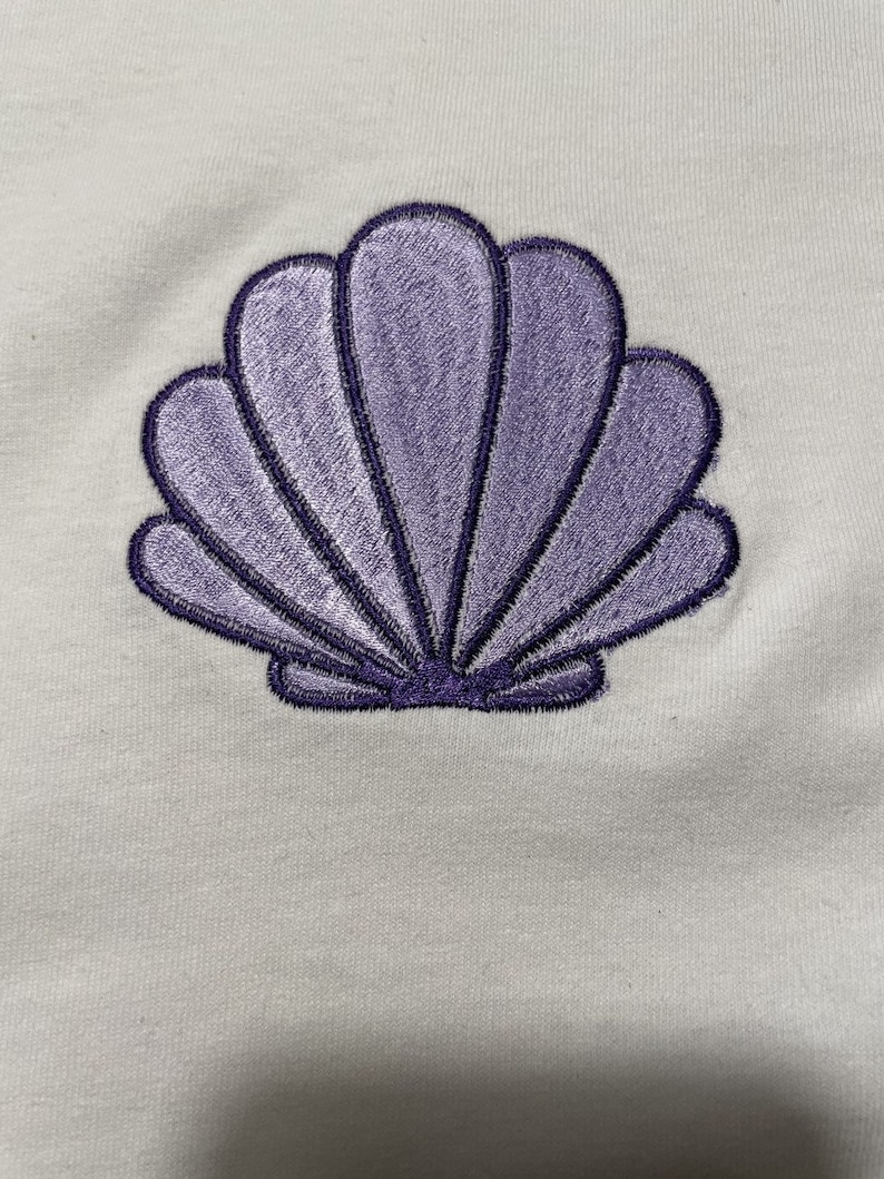 May include: A light-colored fabric with a detailed embroidered seashell design. The seashell is a soft purple color with darker purple outlining, and the design is centered on the fabric. The seashell has a fan-like shape with multiple curved sections.