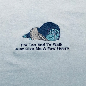 May include: Light blue t-shirt with an embroidered image of a blue character with glasses and the text "I'm Too Sad To Walk Just Give Me A Few Hours."