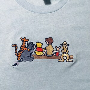 May include: A light blue t-shirt with an embroidered image of Winnie the Pooh and his friends. The characters are Tigger, Piglet, Eeyore, Winnie the Pooh, and Christopher Robin.