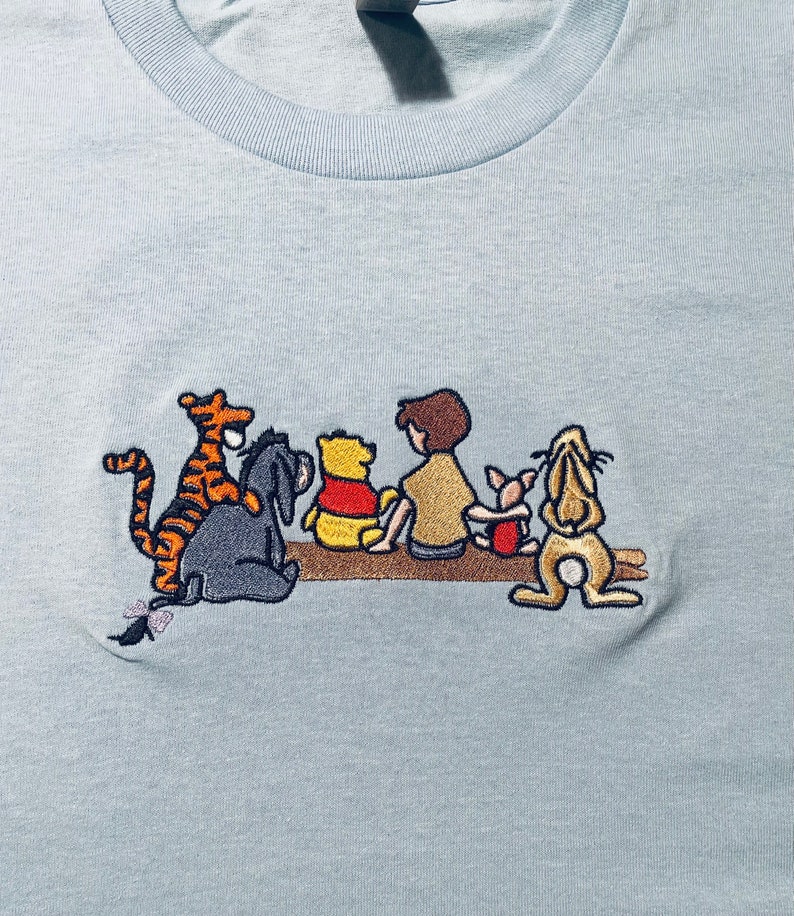 May include: A light blue t-shirt with an embroidered image of Winnie the Pooh and his friends. The characters are Tigger, Piglet, Eeyore, Winnie the Pooh, and Christopher Robin.
