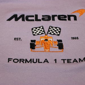 Op de afbeelding: Lavendelkleurig kledingstuk met een geborduurd McLaren Formula 1 Team-ontwerp. Het ontwerp bevat het McLaren-logo, een Formule 1-auto, geblokte vlaggen en de tekst "EST. 1985".