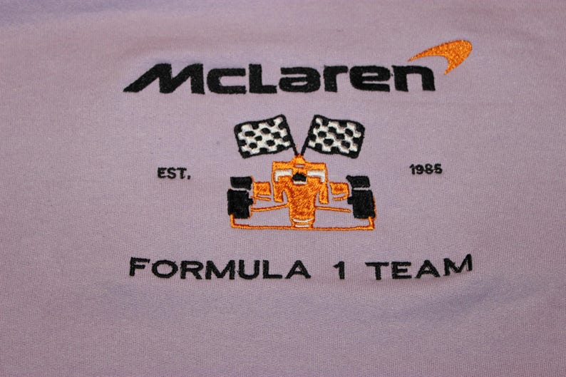 Op de afbeelding: Lavendelkleurig kledingstuk met een geborduurd McLaren Formula 1 Team-ontwerp. Het ontwerp bevat het McLaren-logo, een Formule 1-auto, geblokte vlaggen en de tekst "EST. 1985".