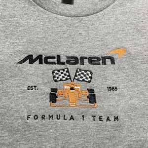 Op de afbeelding: Een grijs T-shirt met ronde hals en het McLaren-logo geborduurd in zwart en oranje. Het ontwerp bevat een Formule 1-auto, geblokte vlaggen en de tekst "EST. 1985 FORMULA 1 TEAM".