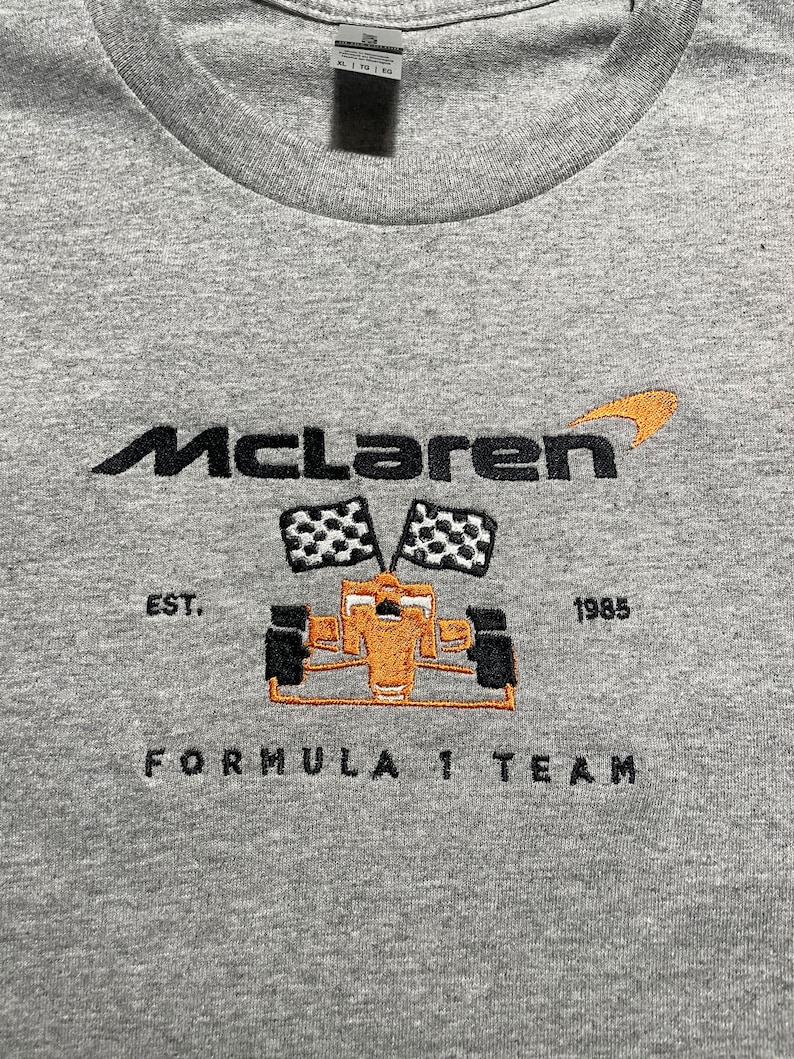 Op de afbeelding: Een grijs T-shirt met ronde hals en het McLaren-logo geborduurd in zwart en oranje. Het ontwerp bevat een Formule 1-auto, geblokte vlaggen en de tekst "EST. 1985 FORMULA 1 TEAM".