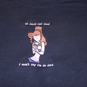May include: A dark blue t-shirt with white embroidered text that reads "at least out loud" and "I won't say I'm in love". The text is centred on the shirt and is surrounded by a cartoon image of a woman with brown hair holding a flower.