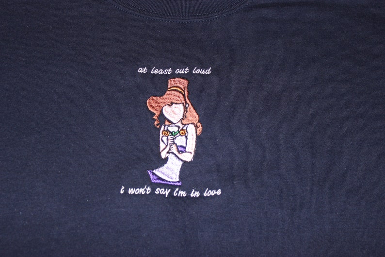 May include: A dark blue t-shirt with white embroidered text that reads "at least out loud" and "I won't say I'm in love". The text is centred on the shirt and is surrounded by a cartoon image of a woman with brown hair holding a flower.