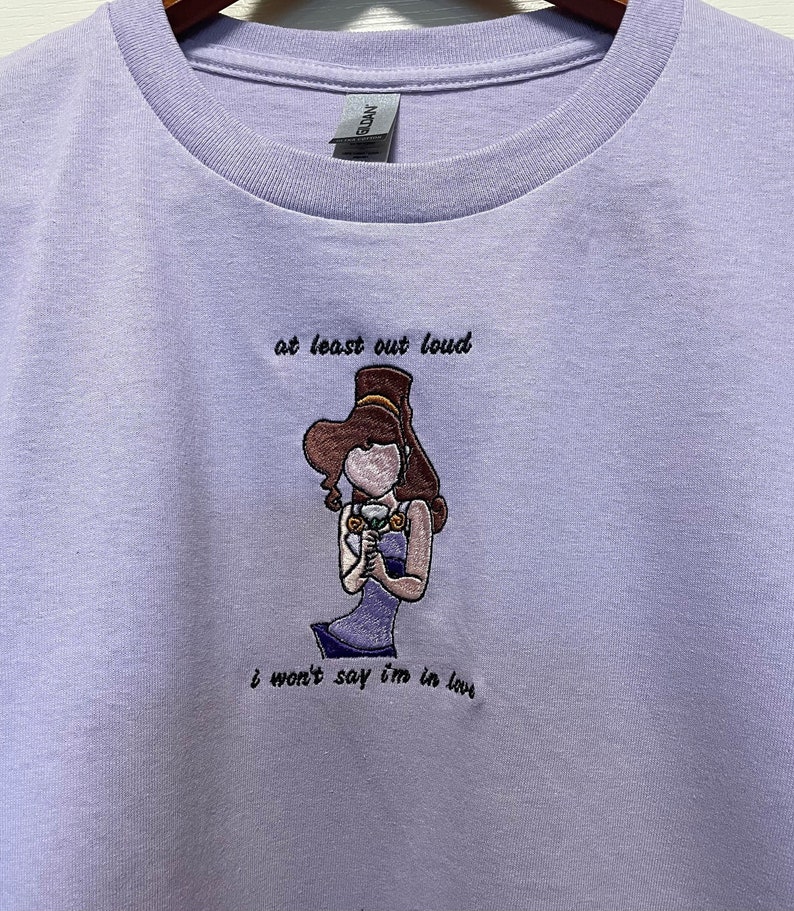Może przedstawiać: Koszulka w kolorze lawendowym z haftowanym obrazem kobiety trzymającej kwiat. Tekst "at least out loud" i "I won't say I'm in love" jest wyszyty pod obrazem.