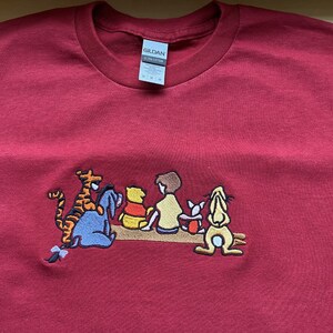 May include: Red t-shirt with an embroidered image of Winnie the Pooh and his friends, including Tigger, Piglet, Eeyore, and Rabbit. The characters are sitting on a log.