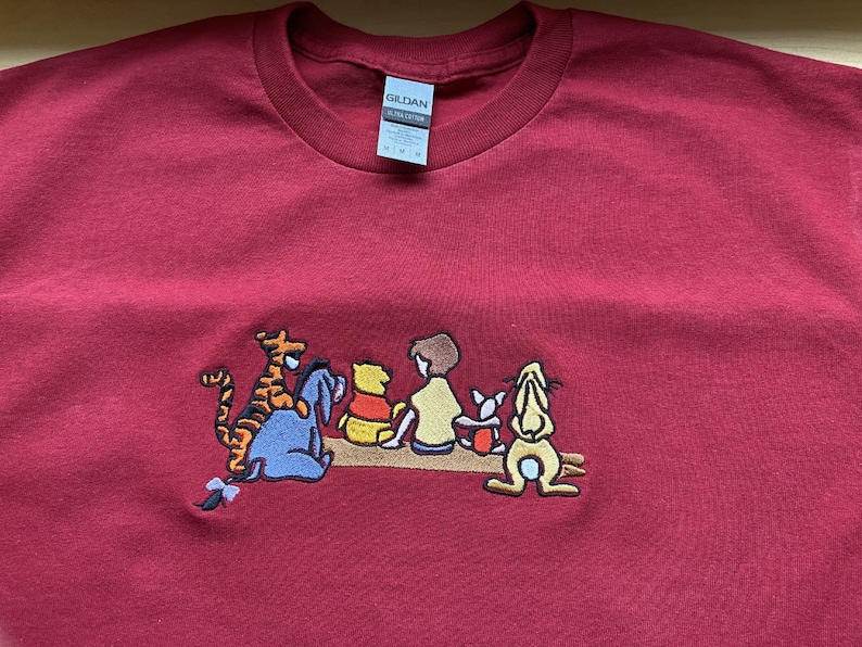 May include: Red t-shirt with an embroidered image of Winnie the Pooh and his friends, including Tigger, Piglet, Eeyore, and Rabbit. The characters are sitting on a log.