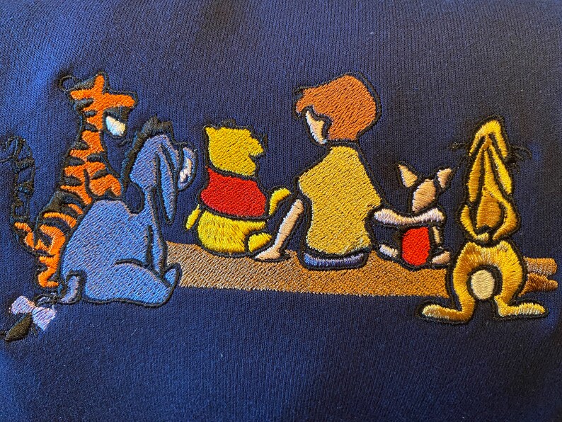 May include: Embroidered Winnie the Pooh and friends design on a navy blue sweatshirt. The characters include Winnie the Pooh, Piglet, Eeyore, Tigger, and Christopher Robin.