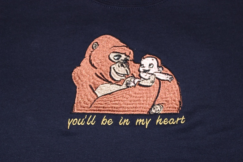Puede incluir: Una camiseta azul marino con una imagen bordada de un orangut&aacute;n sosteniendo a un beb&eacute; orangut&aacute;n. El texto "you'll be in my heart" est&aacute; bordado en amarillo debajo de la imagen.