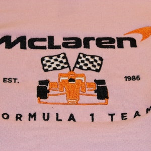 Op de afbeelding: Zachtroze sweatshirt met een geborduurd McLaren Formula 1 Team logo. Het logo toont de naam McLaren in zwart, een oranje swoosh, een raceauto, geblokte vlaggen en de tekst "EST. 1985".