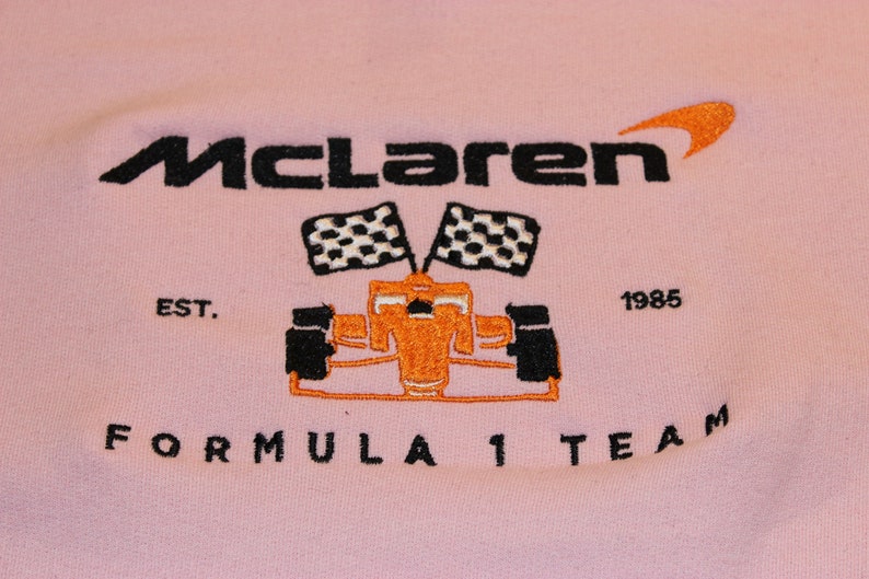 Op de afbeelding: Zachtroze sweatshirt met een geborduurd McLaren Formula 1 Team logo. Het logo toont de naam McLaren in zwart, een oranje swoosh, een raceauto, geblokte vlaggen en de tekst "EST. 1985".