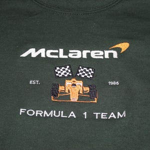 Op de afbeelding: Donkergroene sweatshirt met geborduurd McLaren Formula 1 Team logo. Het logo bevat de naam McLaren in het wit, een gestileerde oranje swoosh, een Formule 1-auto en geblokte vlaggen. De tekst "EST. 1985" is ook inbegrepen.