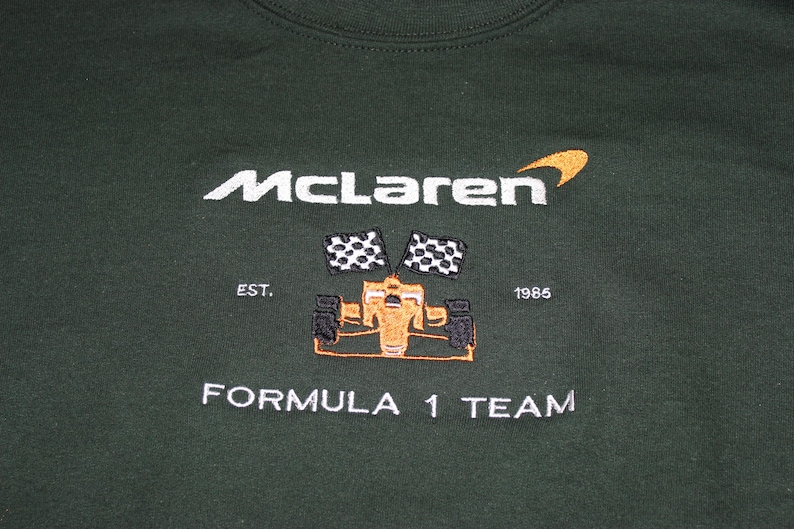 Op de afbeelding: Donkergroene sweatshirt met geborduurd McLaren Formula 1 Team logo. Het logo bevat de naam McLaren in het wit, een gestileerde oranje swoosh, een Formule 1-auto en geblokte vlaggen. De tekst "EST. 1985" is ook inbegrepen.