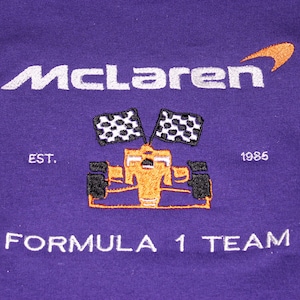 Op de afbeelding: Paarse stof met geborduurd McLaren Formula 1 Team logo. Het logo bevat de naam McLaren in wit, een oranje swoosh, een Formule 1-auto, twee geblokte vlaggen en de tekst "EST. 1985".