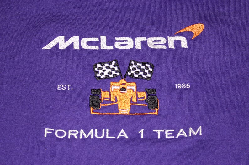 Op de afbeelding: Paarse stof met geborduurd McLaren Formula 1 Team logo. Het logo bevat de naam McLaren in wit, een oranje swoosh, een Formule 1-auto, twee geblokte vlaggen en de tekst "EST. 1985".