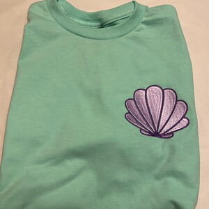 May include: A folded, light green t-shirt with a purple seashell embroidered on the front. The t-shirt has a crew neck and is made of a soft material. The seashell design is detailed with purple thread.