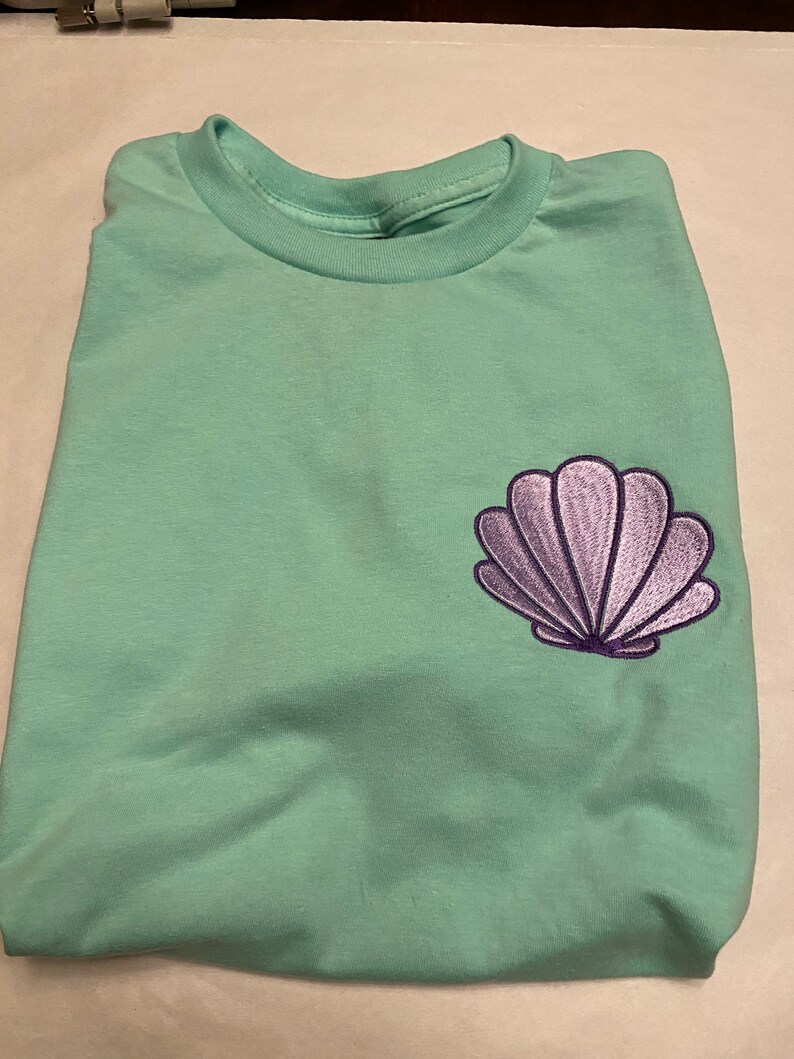May include: A folded, light green t-shirt with a purple seashell embroidered on the front. The t-shirt has a crew neck and is made of a soft material. The seashell design is detailed with purple thread.
