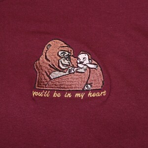 Puede incluir: Una camiseta de color granate con una imagen bordada de un orangut&aacute;n sosteniendo a un beb&eacute; orangut&aacute;n. El texto "you'll be in my heart" est&aacute; bordado debajo de la imagen.