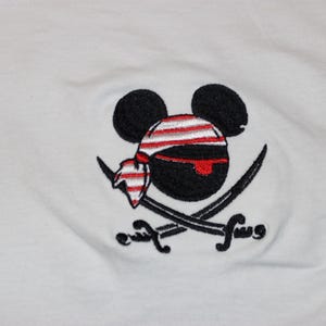 May include: White t-shirt with an embroidered pirate-themed design. The design features a black mouse head wearing a red and white striped bandana, with two crossed swords. The design is centrally placed.