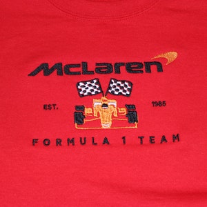 Op de afbeelding: Rode sweatshirt met geborduurd McLaren Formula 1 Team logo. Het logo bevat de naam McLaren, een gestileerde oranje swoosh, een raceauto en geblokte vlaggen. De tekst "EST. 1985" is ook geborduurd.