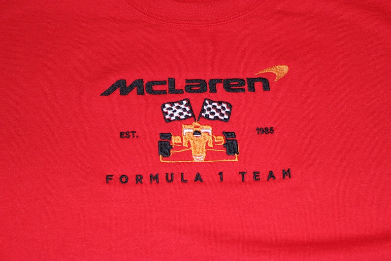 Op de afbeelding: Rode sweatshirt met geborduurd McLaren Formula 1 Team logo. Het logo bevat de naam McLaren, een gestileerde oranje swoosh, een raceauto en geblokte vlaggen. De tekst "EST. 1985" is ook geborduurd.
