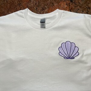 May include: White t-shirt with a small, embroidered seashell design in shades of purple. The seashell is centered on the left chest area of the shirt. The shirt is made of a soft, likely cotton material.