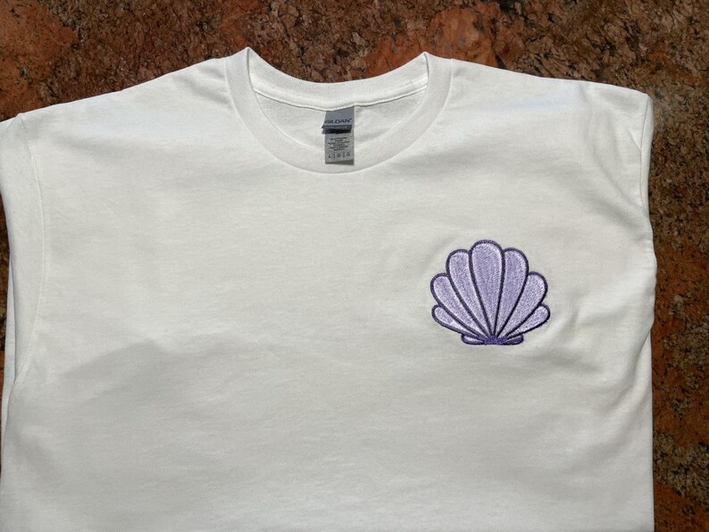 May include: White t-shirt with a small, embroidered seashell design in shades of purple. The seashell is centered on the left chest area of the shirt. The shirt is made of a soft, likely cotton material.