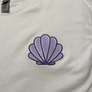May include: White t-shirt with a purple seashell embroidery on the front. The seashell design is detailed with a dark purple outline and shading, creating a three-dimensional effect. The shirt appears to be made of a soft, comfortable material.