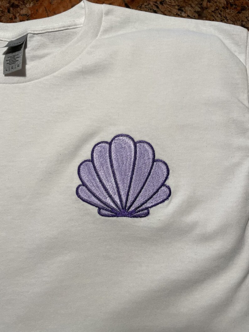May include: White t-shirt with a purple seashell embroidery on the front. The seashell design is detailed with a dark purple outline and shading, creating a three-dimensional effect. The shirt appears to be made of a soft, comfortable material.
