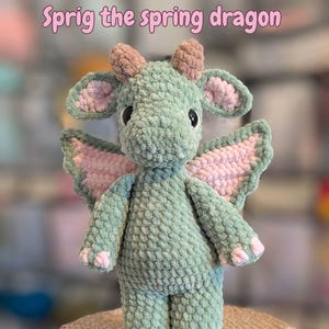 May include: A crocheted dragon toy in green and pink. The dragon has large eyes, small horns, and wings. The text "Sprig the spring dragon" is written above the dragon in pink letters. The dragon is sitting on a woven surface.