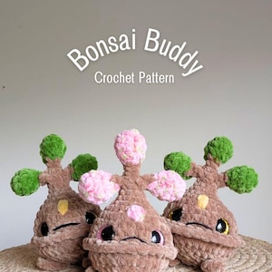 May include: Three plush crochet figures labeled Bonsai Buddy Crochet Pattern. Two figures have brown bodies, green foliage, and yellow accents. The center figure has a brown body with pink blossoms. All have large dark eyes and small feet.