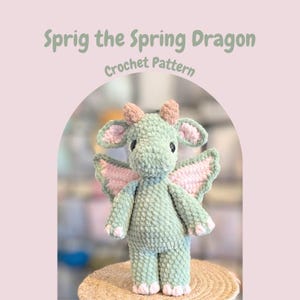 May include: A crocheted dragon toy in shades of green, pink, and white. The dragon has wings, horns, and a friendly expression. The text "Sprig the Spring Dragon Crochet Pattern" is above the dragon.