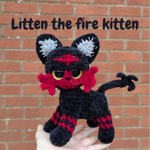 May include: A handmade crochet Litten the fire kitten plush toy. The toy is black with red and gold details. The text "Litten the fire kitten" is above the toy. The kitten has a red face and paws, with a red marking on its head.