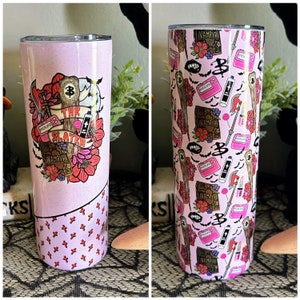 May include: A pink stainless steel tumbler with a graphic of a vampire slayer themed design. The design includes a stake, a book, a charm bracelet, and a cross. The tumbler has a pink and white cross pattern on the bottom half.