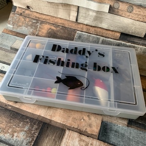 May include: A clear plastic tackle box with multiple compartments. The lid has a black vinyl sticker that says "Daddy's Fishing Box" with a black silhouette of a fish and a fishing hook.