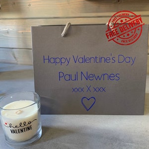 May include: A grey gift bag with the text "Happy Valentine's Day Paul Newnes XXX XXXX" and a heart. A small candle with the text "hello VALENTINE" is in the foreground.