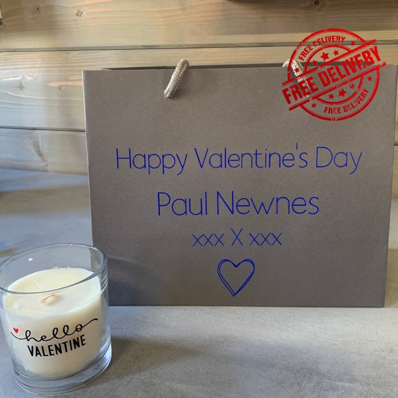 May include: A grey gift bag with the text "Happy Valentine's Day Paul Newnes XXX XXXX" and a heart. A small candle with the text "hello VALENTINE" is in the foreground.