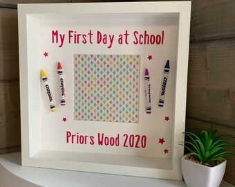 May include: White picture frame with a pink background and the text "My First Day at School" and "Priors Wood 2020". The frame has a picture of a colorful patterned paper inside. There are four crayons on the frame, two on each side of the picture.