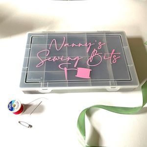 May include: A clear plastic storage box with a black lid. The lid has a pink vinyl sticker that says "Nanny's Sewing Bits" with a pink needle and thread illustration.