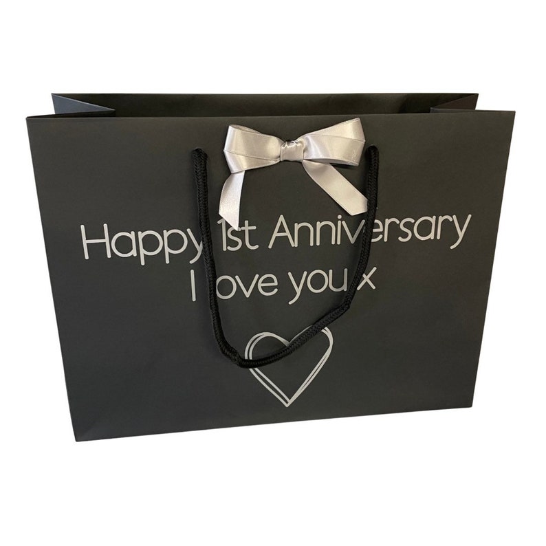 May include: Black gift bag with a silver ribbon and the text "Happy 1st Anniversary I love you x" printed on the front. A silver heart is printed below the text.