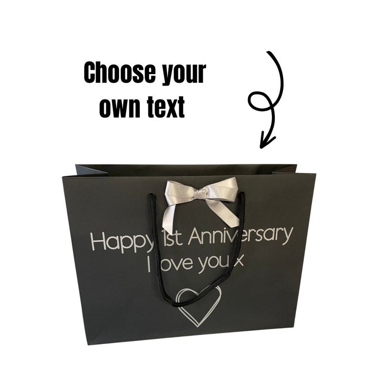 May include: A black gift bag with a white satin bow and black handles. The bag has the text "Happy 1st Anniversary I love you x" printed on it in silver lettering. There is a silver heart design at the bottom of the bag.