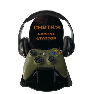 May include: A black gaming headset with a green Xbox controller on a black stand. The stand has the text "CHRIS'S GAMING STATION" in orange pixelated letters.