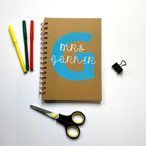 May include: A brown spiral notebook with a blue letter G on the front. The text "mrs garner" is written in white on the blue letter G.