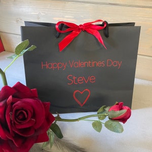 May include: Black gift bag with a red ribbon and the text "Happy Valentines Day Steve" and a red heart.