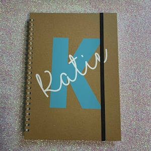 May include: A brown spiral bound notebook with a blue letter "K" and the name "Katin" written in white cursive on the front cover.