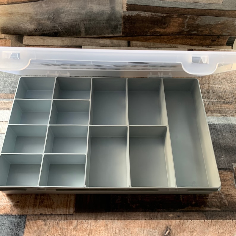 May include: A clear plastic storage box with a gray interior. The box has multiple compartments of varying sizes, perfect for organizing small items.