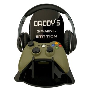 May include: Black gaming station with the text "DADDY'S GAMING STATION" in white. It holds a green Xbox controller and black headphones. The station is designed to organize gaming accessories.